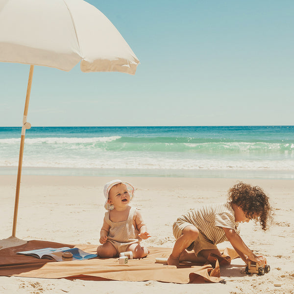 a toddler and a baby are sitting on the beach blanket on the beach, playing their toys.