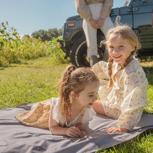 One girl is laying on the picnic blanket and one girl is siting  on the picnic blanket and smiling.  The mum is leaning on the car and watching their play at the back.