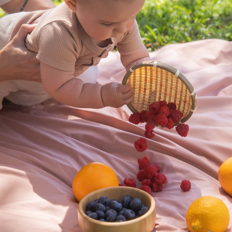 a baby is having a picnic on the picnic blanket and pulling all the raspberries on blanket.  Some oranges and blueberries are placed on picnic blanket. 